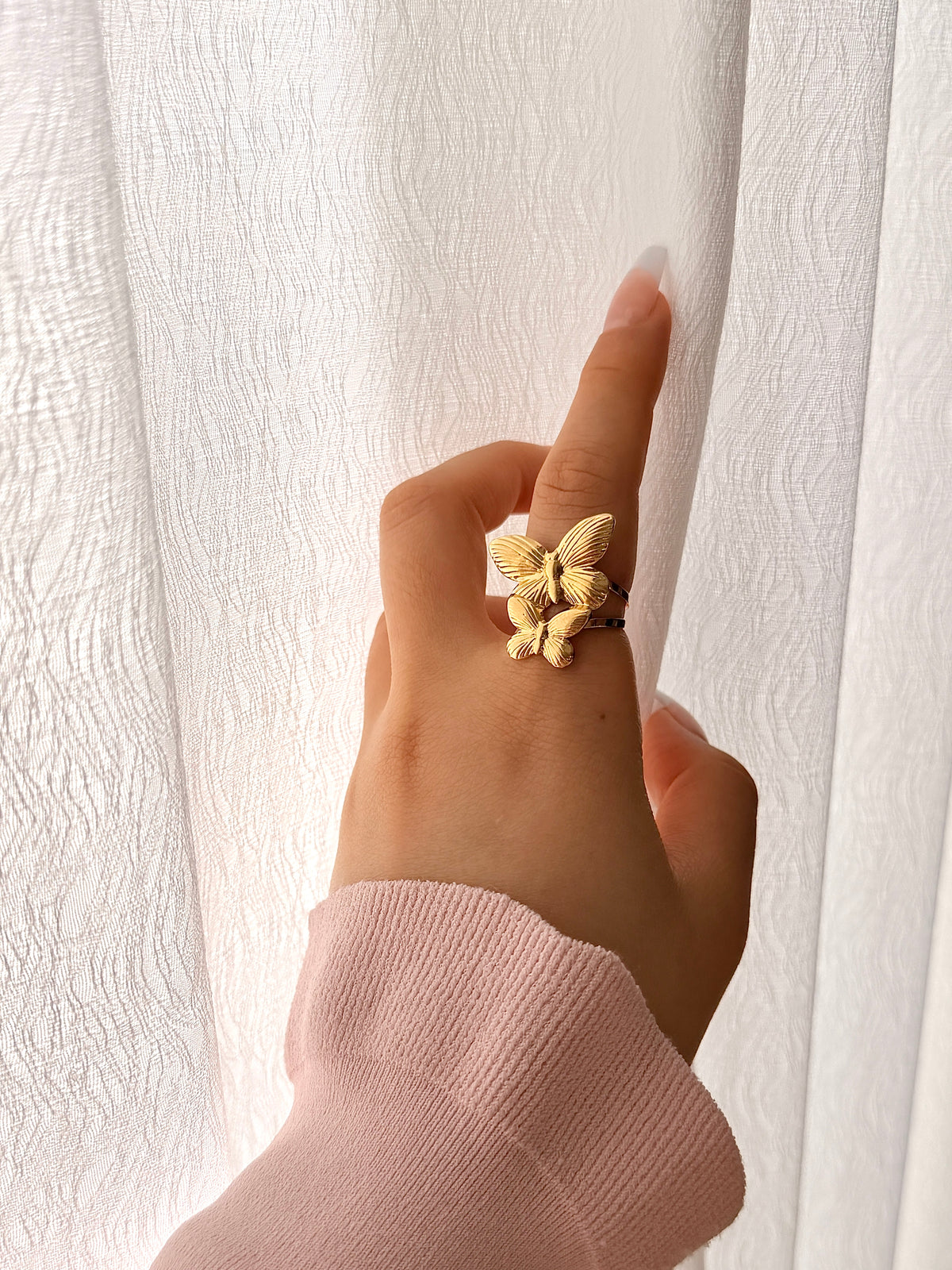 Hand wearing a gold butterfly ring against a white curtain background