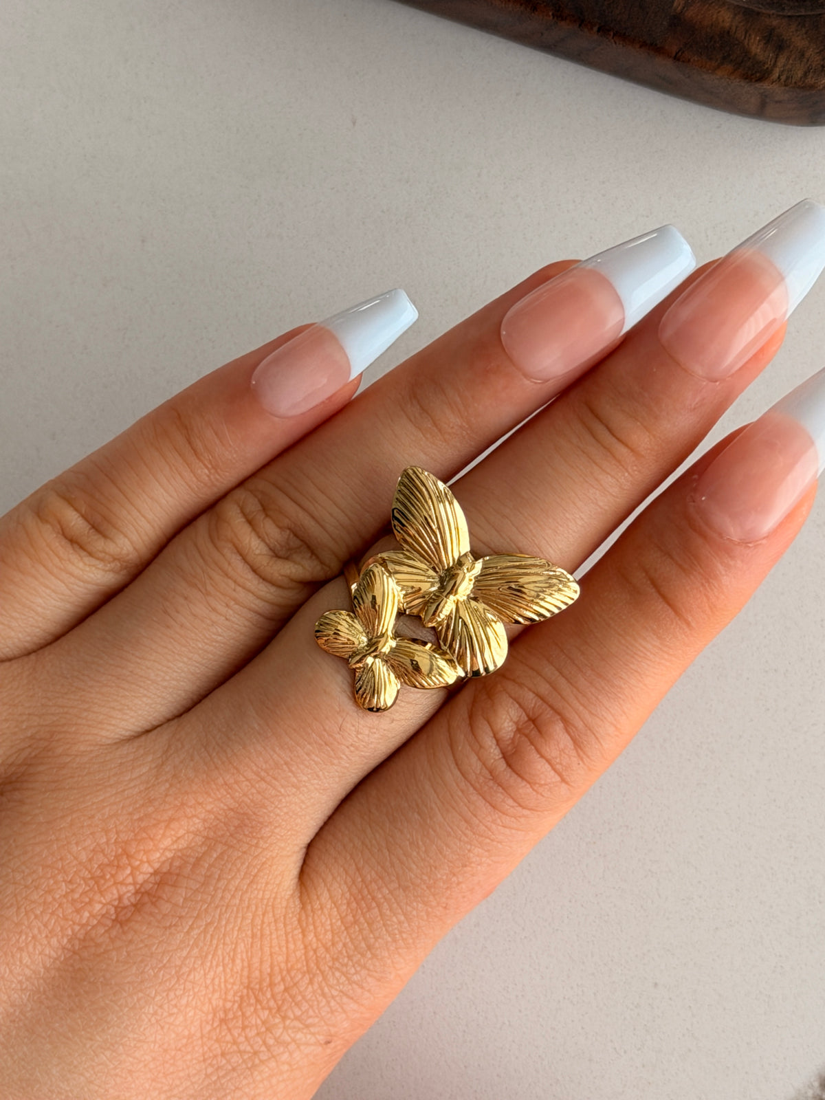 Gold butterfly ring on a hand with white nail polish against a neutral background